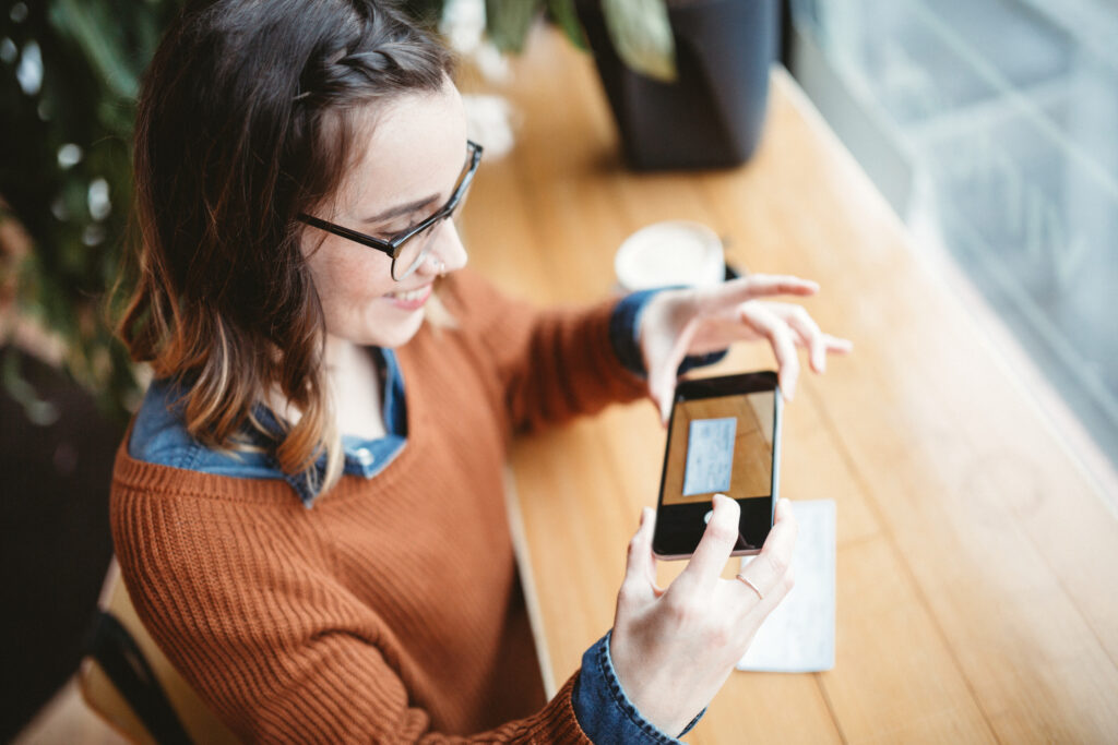 A smiling young woman takes a picture with her smart phone of a check or paycheck for digital electronic depositing, also known as "Remote Deposit Capture".  She sits in a coffee shop, enjoying an espresso latte.  Bright sunlight shines in the window.  Horizontal image.