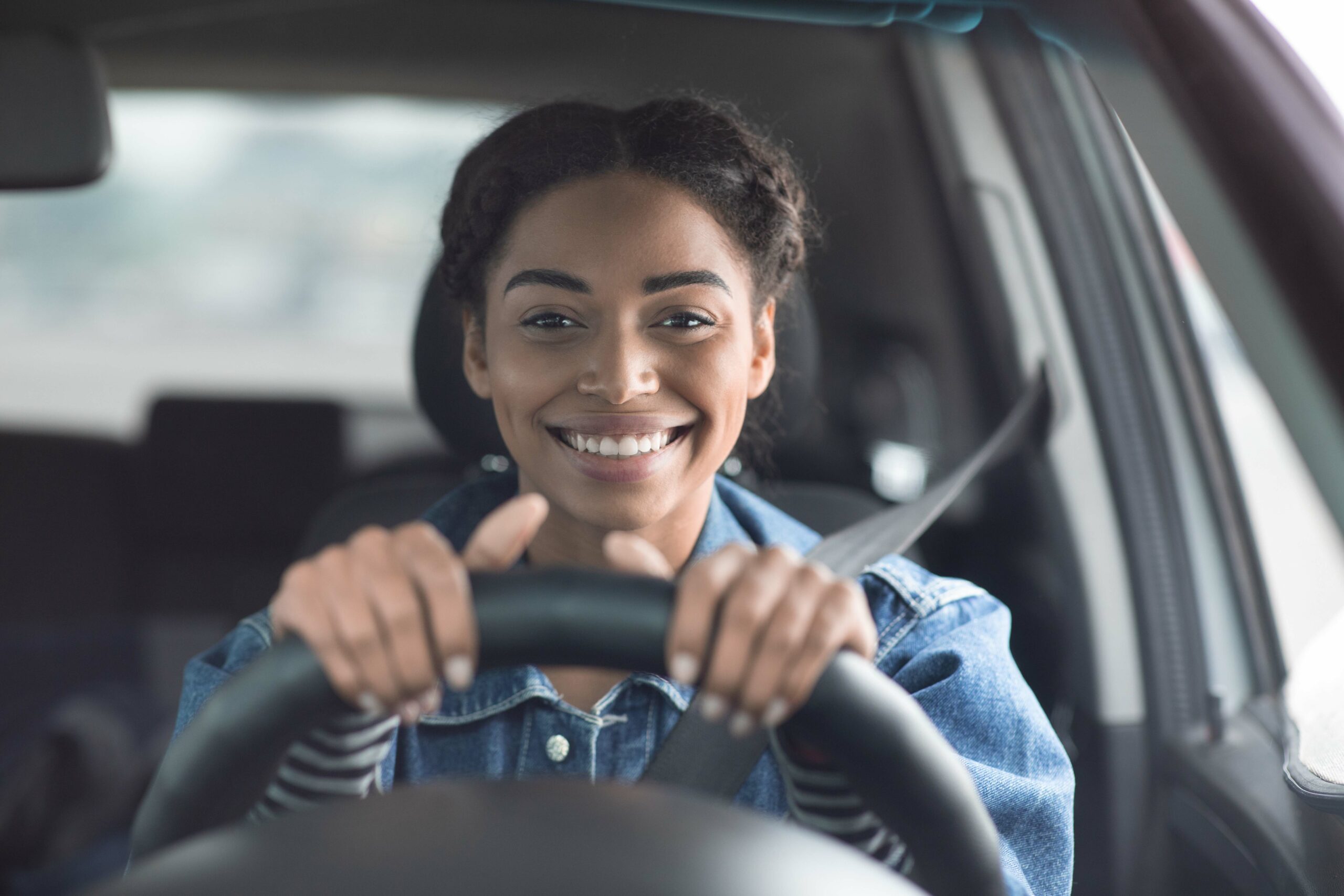 Road to vacation and traveling in new city. Cheerful millennial african american lady with fastened belt drives car, holds wheel and looks through windshield going to journey or work on taxi in town